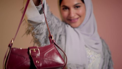 شراء بضائع الماركات العالمية، متابعة آخر صيحات الموضة, الاهتمام بالمظهر الخارجي، تصوير مقرب لامرأة عربية خليجية اماراتية ترتدي الحجاب والعباءة تقف باستقامة بتعابير الثقة بالنفس، سيدة سعودية تنظر الى الكاميرا وتحمل بيدها حقيبة حمراء، خلفية ملونة
