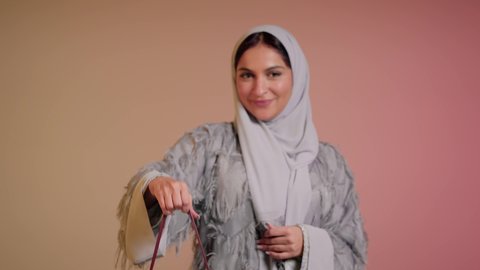 متابعة آخر صيحات الموضة, الاهتمام بالمظهر الخارجي، شراء بضائع الماركات العالمية، تصوير مقرب لامرأة عربية خليجية اماراتية ترتدي الحجاب والعباءة تقف باستقامة بتعابير الثقة بالنفس، سيدة سعودية تنظر الى الكاميرا وتحمل بيدها حقيبة حمراء، خلفية ملونة