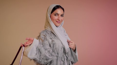 متابعة آخر صيحات الموضة, الاهتمام بالمظهر الخارجي، شراء بضائع الماركات العالمية، تصوير مقرب لامرأة عربية خليجية اماراتية ترتدي الحجاب والعباءة تقف باستقامة بتعابير الثقة بالنفس، سيدة سعودية تنظر الى الكاميرا وتحمل بيدها حقيبة حمراء، خلفية ملونة