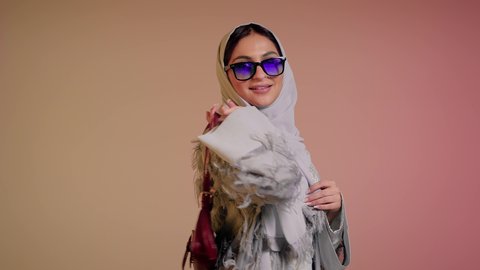 متابعة آخر صيحات الموضة, شراء بضائع الماركات العالمية، الاهتمام بالمظهر الخارجي، تصوير مقرب لامرأة عربية خليجية اماراتية ترتدي الحجاب والعباءة مع النظارات الشمسية تقف باستقامة بتعابير الثقة بالنفس، سيدة سعودية تنظر الى الكاميرا وتحمل بيدها حقيبة حمراء، خلفية ملونة