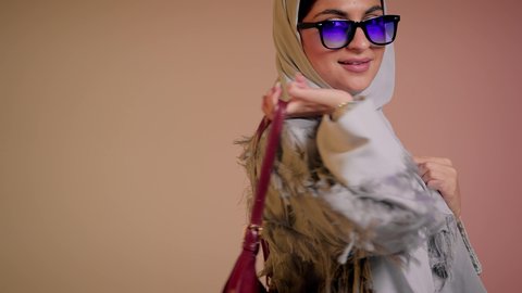 شراء بضائع الماركات العالمية، متابعة آخر صيحات الموضة, الاهتمام بالمظهر الخارجي، تصوير مقرب لامرأة عربية خليجية اماراتية ترتدي الحجاب والعباءة مع النظارات الشمسية تقف باستقامة بتعابير الثقة بالنفس، سيدة سعودية تنظر الى الكاميرا وتحمل بيدها حقيبة حمراء، خلفية ملونة