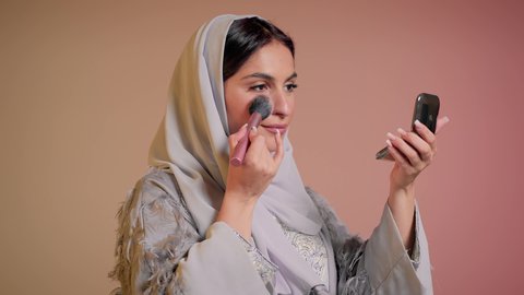 التسويق لمستحضرات ومنتجات التجميل, الاناقة والاهتمام بالمظهر الخارجي, استخدام المساحيق التجميلية, مفهوم التزين والتجمل, تصوير مقرب لامرأة عربية خليجية إماراتية محجبة ترتدي العباءة تستعمل فرشاة أحمر الخدود, شابة سعودية تضع المكياج, خلفية ملونة