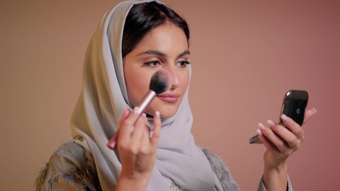 الاهتمام والعناية بالمظهر الخارجي, التسويق لمستحضرات ومنتجات التجميل, استخدام المساحيق التجميلية, مفهوم التزين والتجمل, تصوير مقرب لامرأة عربية خليجية إماراتية محجبة ترتدي العباءة تستعمل فرشاة أحمر الخدود, شابة سعودية تضع المكياج, خلفية ملونة