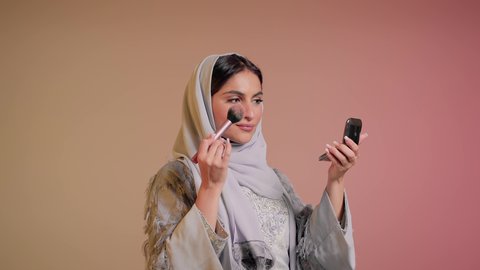 الاهتمام والعناية بالمظهر الخارجي, التسويق لمستحضرات ومنتجات التجميل, استخدام المساحيق التجميلية, مفهوم التزين والتجمل, تصوير مقرب لامرأة عربية خليجية إماراتية محجبة ترتدي العباءة تستعمل فرشاة أحمر الخدود, شابة سعودية تضع المكياج, خلفية ملونة
