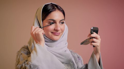 العناية والاهتمام بالمظهر الخارجي، مفهوم التزين والتجمل، ايماءات السعادة والسرور، شابة سعودية تضع المكياج, الترويج لمنتجات التجميل, استخدام المستحضرات التجميلية, تصوير مقرب لامرأة عربية خليجية إماراتية محجبة ترتدي العباءة تقوم باستعمال المسكرة, خلفية ملونة