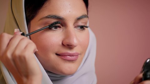 الاهتمام والعناية بالمظهر الخارجي، مفهوم التزين والتجمل، ايماءات السعادة والسرور، شابة سعودية تضع المكياج, الترويج لمنتجات التجميل, استخدام المستحضرات التجميلية, تصوير مقرب لامرأة عربية خليجية إماراتية محجبة ترتدي العباءة تقوم باستعمال المسكرة, خلفية ملونة