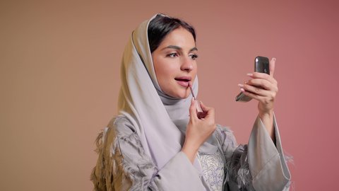 شابة سعودية تقوم بوضع المكياج, العناية بالجمال والمظهر الخارجي, استعمال المستحضرات التجميلية, الترويج لمنتجات و مساحيق التجميل, مفهوم التزين والتجمل,  تصوير مقرب لامرأة عربية خليجية إماراتية محجبة ترتدي العباءة تضع أحمر شفاه, خلفية ملونة