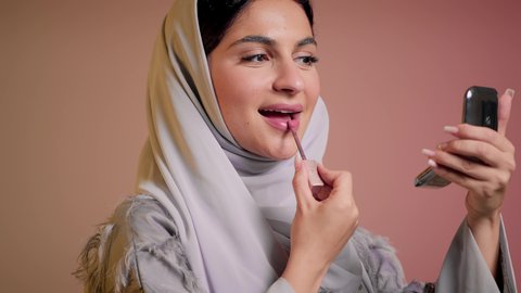 العناية بالجمال والمظهر الخارجي, استعمال المستحضرات التجميلية, الترويج لمنتجات و مساحيق التجميل, مفهوم التزين والتجمل,  تصوير مقرب لامرأة عربية خليجية إماراتية محجبة ترتدي العباءة تضع أحمر شفاه, شابة سعودية تقوم بوضع المكياج, خلفية ملونة