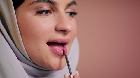 استعمال المستحضرات التجميلية, الترويج لمنتجات و مساحيق التجميل, مفهوم التزين والتجمل, العناية بالجمال والمظهر الخارجي, تصوير مقرب لامرأة عربية خليجية إماراتية محجبة ترتدي العباءة تضع أحمر شفاه, شابة سعودية تقوم بوضع المكياج, خلفية ملونة