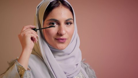 التزين والتجمل والاهتمام بالمظهر الخارجي، ايماءات السعادة والسرور، شابة سعودية تضع المكياج, الترويج لمنتجات التجميل, استخدام المستحضرات التجميلية, تصوير مقرب لامرأة عربية خليجية إماراتية محجبة ترتدي العباءة تقوم باستعمال المسكرة, خلفية ملونة