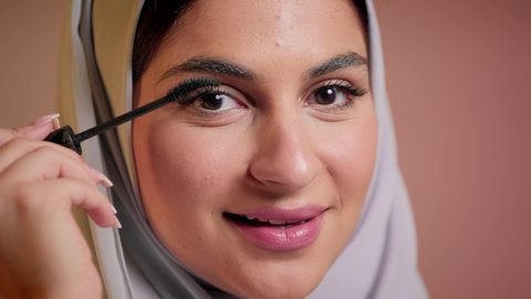 ايماءات السعادة والسرور، شابة سعودية تضع المكياج, الترويج لمنتجات التجميل, استخدام المستحضرات التجميلية, الاهتمام بالجمال والمظهر الخارجي, مفهوم التزين والتجمل, تصوير مقرب لامرأة عربية خليجية إماراتية محجبة ترتدي العباءة تقوم باستعمال المسكرة, خلفية ملونة