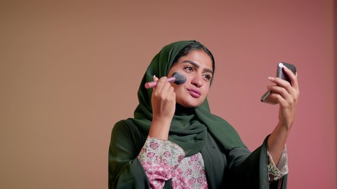 الاهتمام والعناية بالمظهر الخارجي, التسويق لمستحضرات ومنتجات التجميل, استخدام المساحيق التجميلية, مفهوم التزين والتجمل, تصوير مقرب لامرأة عربية خليجية إماراتية محجبة ترتدي العباءة تستعمل فرشاة أحمر الخدود, شابة سعودية تضع المكياج, خلفية ملونة
