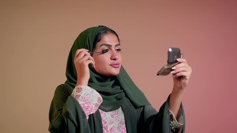 شابة سعودية تضع المكياج, الترويج لمنتجات التجميل, استخدام المستحضرات التجميلية, الشغف بوضع الميك اب، الاهتمام بالجمال والمظهر الخارجي, مفهوم التزين والتجمل, تصوير مقرب لامرأة عربية خليجية إماراتية محجبة ترتدي العباءة تقوم باستعمال المسكرة, خلفية ملونة