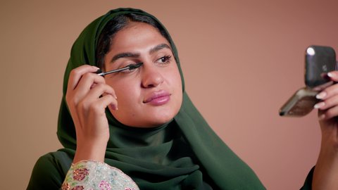 الترويج لمنتجات التجميل, استخدام المستحضرات التجميلية, الشغف بوضع الميك اب، الاهتمام بالجمال والمظهر الخارجي, مفهوم التزين والتجمل, تصوير مقرب لامرأة عربية خليجية إماراتية محجبة ترتدي العباءة تقوم باستعمال المسكرة, شابة سعودية تضع المكياج, خلفية ملونة