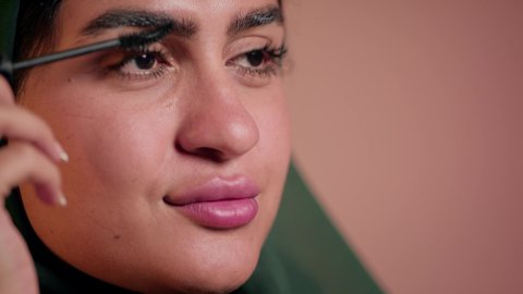 استخدام المستحضرات التجميلية, الشغف بوضع الميك اب، الاهتمام بالجمال والمظهر الخارجي, الترويج لمنتجات التجميل, مفهوم التزين والتجمل, تصوير مقرب لامرأة عربية خليجية إماراتية محجبة ترتدي العباءة تقوم باستعمال المسكرة, شابة سعودية تضع المكياج, خلفية ملونة