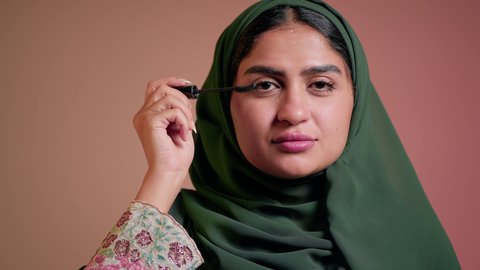 الاهتمام بالجمال والمظهر الخارجي, الترويج لمنتجات ومستحضرات التجميل, مفهوم التزين والتجمل, تصوير مقرب لامرأة عربية خليجية إماراتية محجبة ترتدي العباءة تقوم باستخدام المسكرة, شابة سعودية تضع المكياج, خلفية ملونة