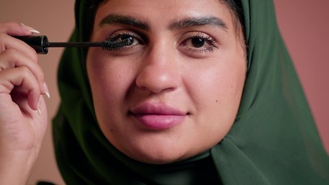 الترويج لمنتجات ومستحضرات التجميل, الاهتمام بالجمال والمظهر الخارجي, مفهوم التزين والتجمل, تصوير مقرب لامرأة عربية خليجية إماراتية محجبة ترتدي العباءة تقوم باستخدام المسكرة, شابة سعودية تضع المكياج, خلفية ملونة