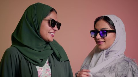 إيماءات الانوثه والدلع والدلال، متابعة أحدث صيحات الموضة، التسويق لماركة ومنتج محلي، بورتريه مقرب لسيدتين عربيتين خليجيتين سعوديتين ترتديان العباءة ونظاره شمسيه ملونه، صديقتان امراتيتان محجبتان تقومان تبادل الأحاديث بتعابير السعادة، خلفية وردية