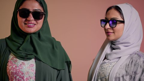 Marketing a local brand and product, interest in outward appearance, keeping up with the latest fashion trends, close-up portrait of two Saudi Arabian Gulf women wearing abaya and colored sunglasses, two veiled female friends looking at the camera with feminine, coquettish and pampering gestures, colored background