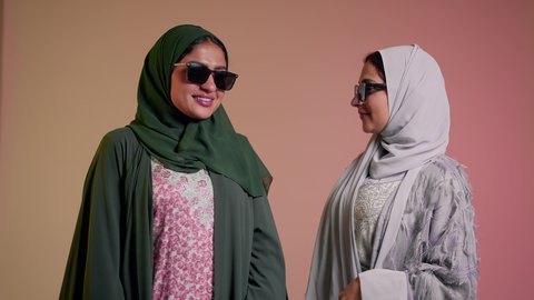 Following the latest fashion trends, marketing a local brand and product, taking care of one's appearance, a close-up portrait of two Saudi Arabian Gulf women wearing an abaya and colored sunglasses, two veiled female friends looking at the camera with feminine gestures, coquettishness, and pampering, a colored background