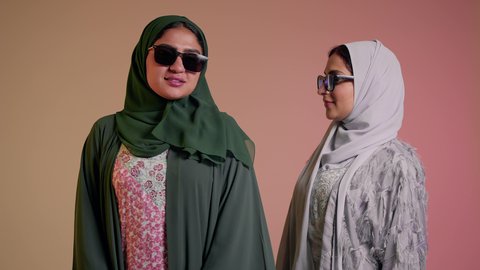 Keeping up with the latest fashion trends, the concept of elegance and attention to one's appearance, a close-up portrait of two Saudi Arabian Gulf women wearing the abaya and colored sunglasses, two veiled female friends looking at the camera with feminine, coquettish and pampering gestures, colored background