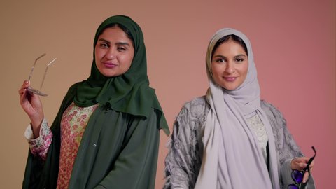 متابعة أحدث صيحات الموضة، التسويق لماركة ومنتج محلي، الاهتمام بالمظهر الخارجي، بورتريه مقرب لسيدتين عربيتين خليجيتين سعوديتين ترتديان العباءة ونظاره شمسيه ملونه، صديقتان امراتيتان محجبتان تنظران إلى الكاميرا بإيماءات الانوثه والدلع والدلال، خلفية ملونة