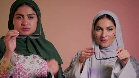 Expressions of self-confidence, following the latest fashion trends, marketing a local brand and product, close-up portrait of two Saudi Arabian Gulf women wearing abaya and colored sunglasses, two veiled female friends looking at the camera with feminine, coquettish and pampering gestures, colored background