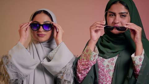 مواكبة أحدث صيحات الموضة والأزياء، مفهوم الأناقة والاهتمام بالظهر الخارجي، بورتريه مقرب لسيدتين عربيتين خليجيتين سعوديتين ترتديان العباءة ونظاره شمسيه ملونه، صديقتان امراتيتان محجبتان تنظران إلى الكاميرا بإيماءات الانوثه والدلع والدلال، خلفية ملونة
