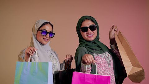 النظر إلى الكاميرا بإيماءات الانوثه والدلال، الأثر الايجابي للتسوق على المرأة، شراء الهدايا والمستلزمات، بورتريه مقرب لسيدتين عربيتين خليجيتين سعوديتين ترتديان العباءة ونظاره شمسيه ملونه، صديقتان امراتيتان محجبتان تحملان أكياس ورقية بتعابير السرور، خلفية وردية