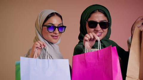 اقتناء الهدايا والمستلزمات، النظر إلى الكاميرا بإيماءات الانوثه والدلال، الاستفادة من العروض والتخفيضات، بورتريه مقرب لسيدتين عربيتين خليجيتين سعوديتين ترتديان العباءة ونظاره شمسيه ملونه، صديقتان امراتيتان محجبتان تحملان أكياس ورقية بتعابير السرور، خلفية وردية