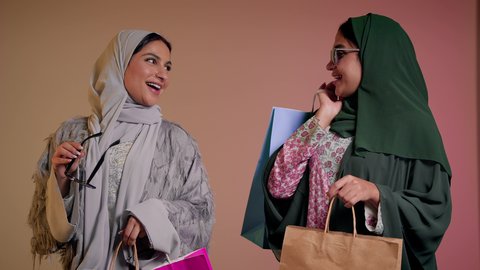 الاستفادة من العروض والتخفيضات الموسمية، اقتناء الهدايا والمستلزمات، النظر إلى الكاميرا بإيماءات الانوثه والدلال، بورتريه لسيدتين عربيتين خليجيتين سعوديتين ترتديان العباءة ونظاره شمسيه ملونه، صديقتان امراتيتان محجبتان تحملان أكياس ورقية بتعابير السرور، خلفية وردية