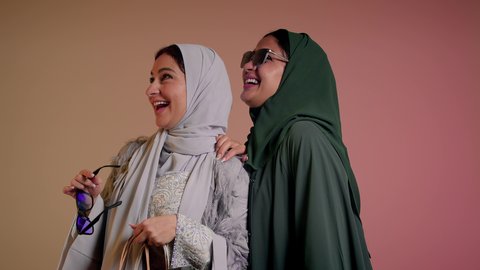 Enjoying shopping, taking advantage of offers and discounts, looking at something with happy gestures, buying gifts and supplies, close-up portrait of two Saudi Arabian Gulf women wearing abaya and tinted sunglasses, two veiled female friends holding paper bags, pink background