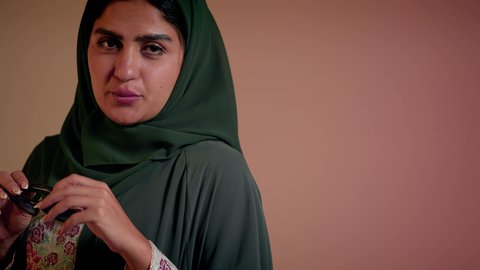 النظر إلى الكاميرا بايماءات السعادة والسرور، قضاء وقت ممتع مع الاصدقاء، مفهوم الأنوثة والأناقة والاهتمام بالمظهر الخارجي، امرأة اماراتية تمسك بيدها نظارة شمسية، بورتريه مقرب لصديقتين عربيتين خليجيتين سعوديتين محجبتين ترتديان العباءة، خلفية وردية