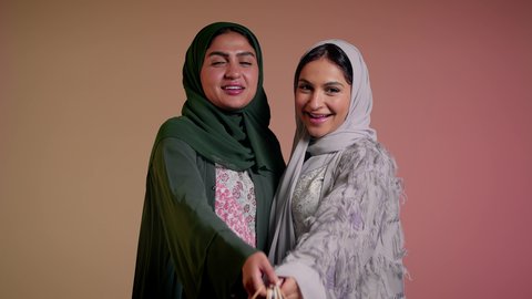 Buying gifts and necessities, enjoying shopping with friends, taking advantage of offers and discounts, looking at the camera with happy gestures, close-up portrait of two Saudi Arabian Gulf women wearing abaya, two veiled female friends holding paper bags, pink background