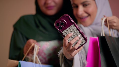 الاستمتاع بالتسوق والتبضع مع الاصدقاء، استخدام الهاتف المحمول، استغلال العروض والحسومات، بورتريه مقرب لسيدة عربية خليجية سعودية تقوم بالتقاط صورة سيلفي تذكارية مع صديقتها بإيماءات السعادة، صديقتان امراتيتان محجبتان ترتديان العباءة تحملان أكياس ورقية، خلفية وردية