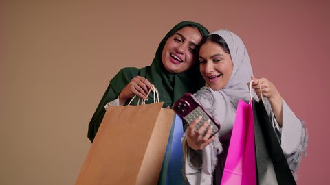 العروض والتخفيضات الموسمية، الاستمتاع بالتسوق والتبضع مع الاصدقاء، استخدام الهاتف المحمول، بورتريه لسيدة عربية خليجية سعودية تقوم بالتقاط صورة سيلفي تذكارية مع صديقتها بإيماءات السعادة والدلع، صديقتان امراتيتان ترتديان العباءة تحملان أكياس ورقية، خلفية وردية
