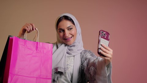 شراء الهدايا والمستلزمات من المحلات والمراكز التجارية، بورتريه مقرب لسيدة عربية خليجية سعودية تقوم بالتقاط صورة سيلفي تذكارية استخدام الهاتف المحمول بإيماءات السعادة والسرور، امرأة اماراتية محجبة ترتدي العباءة تحمل أكياس تسوق ورقية، خلفية وردية