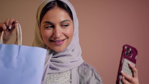 Positive impact of shopping on women, close-up portrait of a Saudi Arabian Gulf woman taking a selfie using a mobile phone with gestures of happiness and pleasure, veiled Emirati woman wearing an abaya holding paper shopping bags, pink background