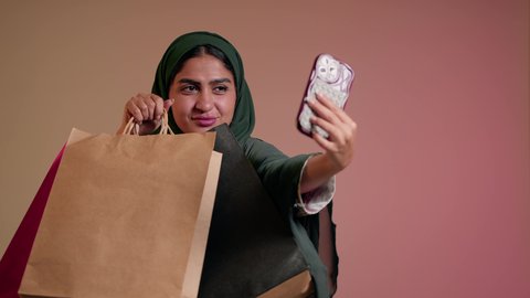 شراء الهدايا والمستلزمات من المحلات والمراكز التجارية، بورتريه مقرب لسيدة عربية خليجية سعودية تقوم بالتقاط صورة سيلفي تذكارية استخدام الهاتف المحمول بإيماءات السعادة والسرور، امرأة اماراتية محجبة ترتدي العباءة تحمل أكياس تسوق ورقية، خلفية وردية