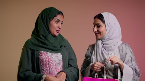 الإشارة بإصبع السبابة إلى شيء ما بإيماءات السعادة، شراء الهدايا والمستلزمات، المتعة بالتسوق والتبضع مع الاصدقاء، استغلال العروض والحسومات، بورتريه لسيدتين عربيتين خليجيتين سعوديتين ترتديان العباءة، صديقتان امراتيتان محجبتان تحملان أكياس ورقية، خلفية وردية