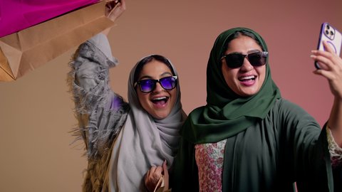 Enjoying shopping with friends, using mobile phone, portrait of a Saudi Arabian Gulf woman taking a selfie with her friend with happy and cheerful gestures, two female friends wearing abaya and sunglasses holding paper bags, pink background