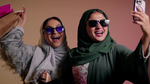 استخدام الهاتف المحمول، الاستمتاع بالتسوق والتبضع مع الاصدقاء، بورتريه لسيدة عربية خليجية سعودية تقوم بالتقاط صورة سيلفي تذكارية مع صديقتها بإيماءات السعادة والمرح، صديقتان امراتيتان ترتديان العباءة وتضعان النظارة الشمسية تحملان أكياس ورقية، خلفية وردية