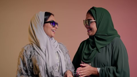 متابعة أحدث صيحات الموضة، التسويق لماركة ومنتج محلي، الاهتمام بالمظهر الخارجي، بورتريه مقرب لسيدتين عربيتين خليجيتين سعوديتين ترتديان العباءة ونظاره شمسيه ملونه، صديقتان امراتيتان محجبتان تنظران إلى الكاميرا بإيماءات الانوثه والدلع والدلال، خلفية ملونة