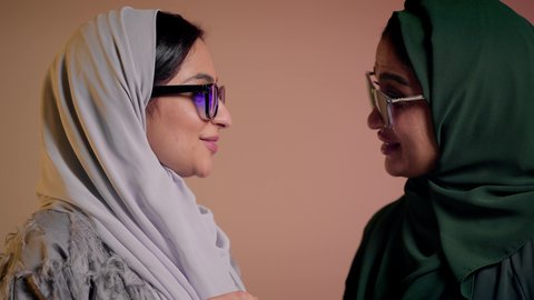 Following the latest fashion trends, marketing a local brand and product, taking care of one's appearance, a close-up portrait of two Saudi Arabian Gulf women wearing an abaya and colored sunglasses, two veiled female friends looking at the camera with feminine gestures, coquettishness, and pampering, a colored background