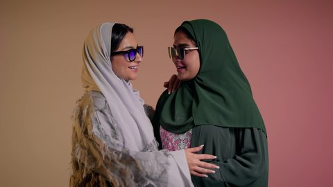 Marketing a local brand and product, keeping up with the latest fashion trends, taking care of one's appearance, a close-up portrait of two Saudi Arabian Gulf women wearing an abaya and colored sunglasses, two veiled female friends looking at the camera with feminine, coquettish and affectionate gestures, a colored background