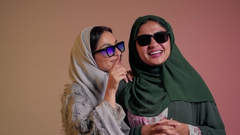 Having fun with friends, elegance and interest in appearance, following the latest fashion trends, close-up portrait of two Saudi Arabian Gulf women wearing abaya and colored sunglasses, two veiled female friends pointing their index fingers at something with happy gestures, colored background