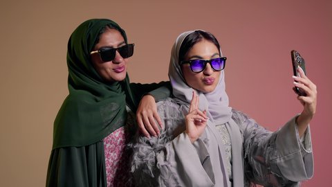 سيدتان عربيتان خليجيتان سعوديتان ترتديان نظاره شمسيه ملونه، صديقتان عربيتان خليجيتان امراتيتان ترتديان العباءه تقومان بالتقاط صور سيلفي تذكاريه عبر الهاتف المحمول, ايماءات  الانوثه والدلال، استخدام احدث التقنيات الحديثه، التطور التقني للاجهزه