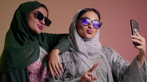 استخدام احدث التقنيات الحديثه، سيدتان عربيتان خليجيتان سعوديتان ترتديان نظاره شمسيه ملونه، صديقتان عربيتان خليجيتان امراتيتان ترتديان العباءه تقومان بالتقاط صور سيلفي تذكاريه عبر الهاتف المحمول بإيماءات  الانوثه والدلع والدلال، خلفية ملونة