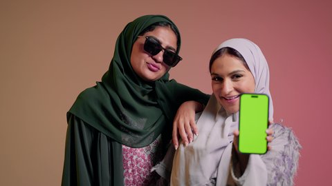 سيده عربيه خليجيه سعوديه تمسك بيدها الهاتف المحمول بشاشه خضراء فارغه برفقه صديقتها، سيدتان عربيتان خليجيتان امراتيتان ترتديان العباءه تنظران الي الكاميرا بثقه وثبات, ايماءات  الانوثه والدلال، كروما