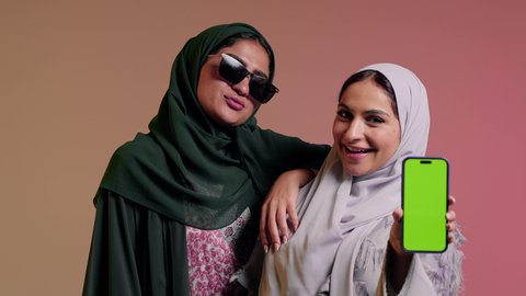 سيده عربيه خليجيه سعوديه تمسك بيدها الهاتف المحمول بشاشه خضراء فارغه برفقه صديقتها، سيدتان عربيتان خليجيتان امراتيتان ترتديان العباءه تنظران الي الكاميرا بثقه وثبات, ايماءات  الانوثه والدلال، كروما